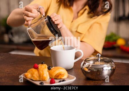 Young female in yellow shirt sitting by table in the kitchen and pouring black tea into cup while going to have breakfast with croissants Stock Photo