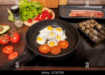 Several fried quail eggs and tomato slices sprinkled with ground black pepper on frying pan surrounded by fresh vegetables Stock Photo