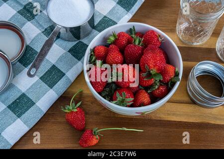 Photograph of freshly picked strawberries in a bowl, getting ready to make strawberry jam, with a cup of sugar, jars, lids, and a towel on a wooden co