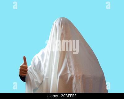 A man covered with a white sheet like a ghost shows a thumb on a blue background Stock Photo
