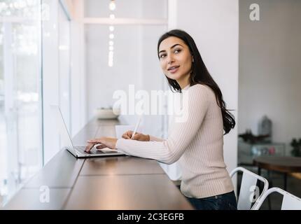 Attractive Indian woman using laptop computer, writing notes, typing on keyboard, working from home. Student learning language, exam preparation Stock Photo