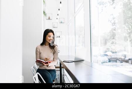 Beautiful Indian woman freelancer using laptop, writing notes, working from home. Asian student studying, exam preparation, distance learning Stock Photo