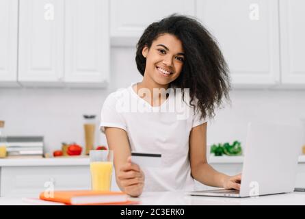 Happy African American woman shopping online, ordering food, standing on kitchen Stock Photo