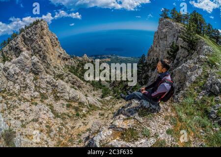 Scenic panoramic high angle view from top of Ai-Petri with a young man in front admiring the mountain view towards coastline between Yalta, Mishor and Stock Photo