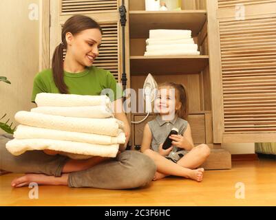 Smiling woman with little daughter sitting near cupboard holding towels in laundry room Stock Photo