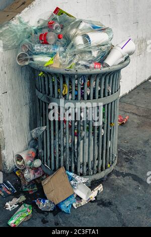 A trash can on a city sidewalk overflows with bottles, cups, food wrappers and more. Stock Photo