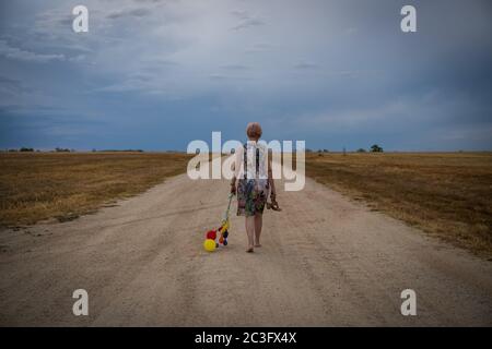 Woman walking barefoot on deserted road carrying shoes and balloons Stock Photo
