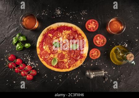 Pepperoni pizza on a black background, with wine glasses and ingredients, tomatoes, basil, and olive oil, shot from the top Stock Photo