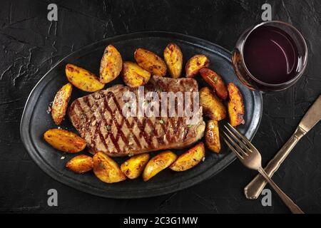 Grilled beef steak with baked potato wedges and red wine, shot from above on a black background Stock Photo