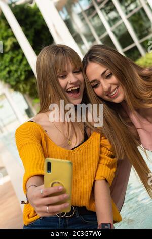 Two young Caucasian girls with blonde hair smile expressively while using a mobile phone sitting on a wooden bench Stock Photo