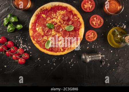 Pepperoni pizza on a black background, with wine glasses and ingredients, tomatoes, basil, and olive oil, shot from above with a Stock Photo