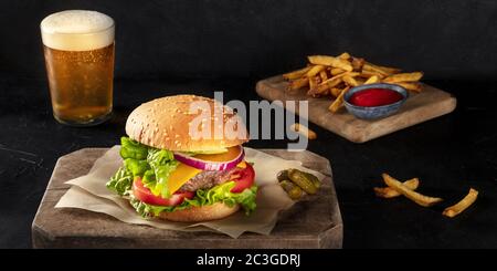 Burger with French fries and beer panorama. Hamburger with beef, cheese, onion, tomato, and green salad, on a dark background. S Stock Photo