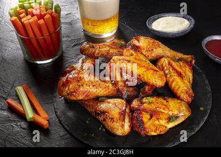 Buffalo chicken wings with cheese and barbecue sauces and celery and carrot sticks, with a glass of beer on a dark background Stock Photo