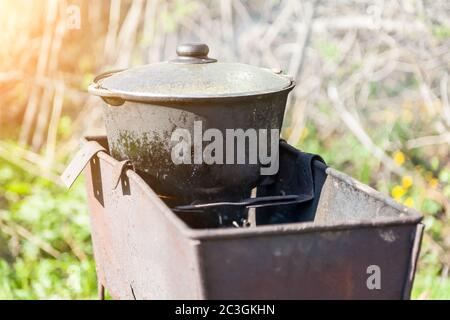 Old traditional national dishes for cooking on an open fire - a cast-iron cauldron inside an iron mongal on coals on a background of green grass in a Stock Photo