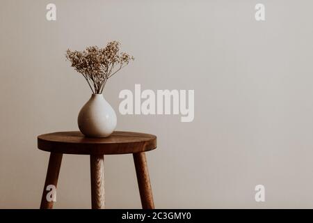 White ceramic vase on a natural brown wooden stool with white dried flowers in an empty room Stock Photo