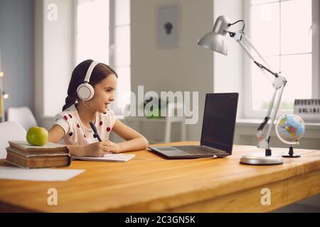 Online school training. Smart girl in headphones is studying a lesson using a laptop sitting at a table at home. Stock Photo
