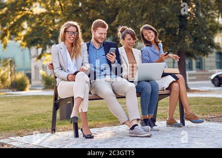 young caucasian businesspeople laughing at park sitting on  a bench with their cell phones, laptop, tablet. outdoor good mood break concept Stock Photo