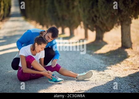 beardy young male personal trainer comfort and advising female who injured her ankle on jogging, because she didn't worm up and stretch before trainin Stock Photo