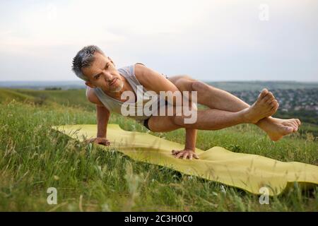 Male yoga on the grass in summer. Mature gray-haired man practicing yoga balance pose in nature Stock Photo
