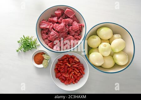 Ingredients for stew or goulash with diced beef, onions, bell pepper, herbs and spices from above on a white painted table, cooking at home during the Stock Photo