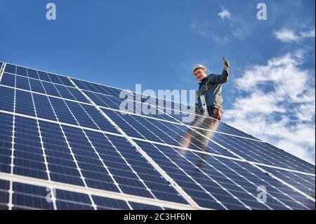 Cheerful male worker giving thumbs up and smiling while installing blue photovoltaic solar panel under beautiful cloudy sky. Electrician showing approval gesture. Concept of alternative energy Stock Photo