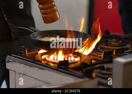 Chef seasoning a pan of food with pepper from a grinder over flames on a gas hob in a kitchen in close up Stock Photo