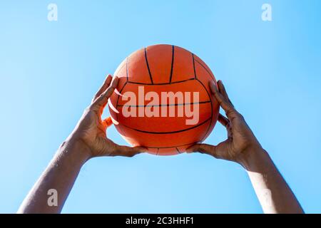 Close up shot of two dark-skinned hands holding up a basketball outdoors against blue clear sky, low angle. Stock Photo