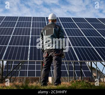 Back view of man electrician in work uniform and safety helmet looking at solar photovoltaic solar panel construction. Concept of alternative energy and power sustainable resources. Stock Photo