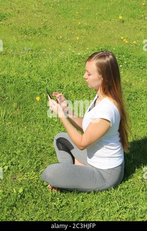 A young beautiful girl of European appearance in a white T-shirt with long blond hair sits on green grass, on the lawn with a mobile phone in her hands, flips through social networks, works in the phone. Stock Photo