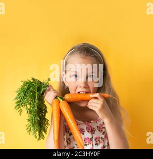 Nice little girl in summer dress with flower pattern and band on hair biting carrot with appetite. Close up shot isolated on yellow