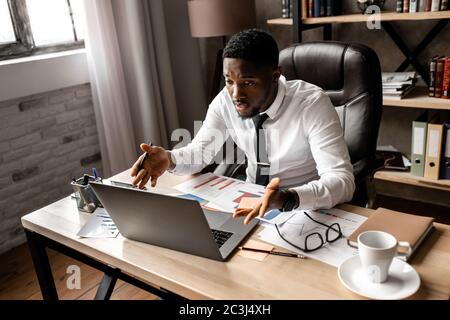 Smiling young african businessman talking to a clients on a video call , sitting at a table in the office Stock Photo