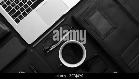 Business desktop concept. Mix of office supplies and gadgets on a black table background. With laptop, mobile phone, glasses, notebook, cup of coffee. Stock Photo