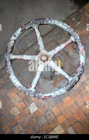 Old rusty valve wheel painted blue red with peeling selective focus with extension device over out of focus background tiled floor and perforated Stock Photo