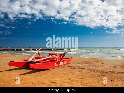Typical red rescue boat, with oars, used by Italian lifeguards, stationary on the sand. "Rescue" writing on the side. Blue sky with white clouds in su Stock Photo