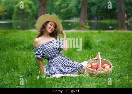 a girl in a hat and a blue dress is sitting on the grass in the Park.girl with a basket of apples near the lilac. Stock Photo