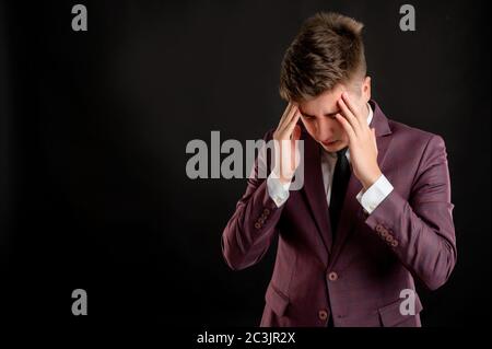 Law student with blond hair dressed in burgundy jacket, white shirt and black tie has headache posing on isolated black background with copy space adv Stock Photo