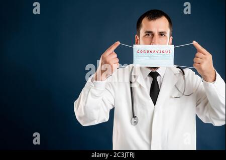 Portrait of male doctor with stethoscope in medical uniform showing protective mask posing on a blue isolated background. Stock Photo