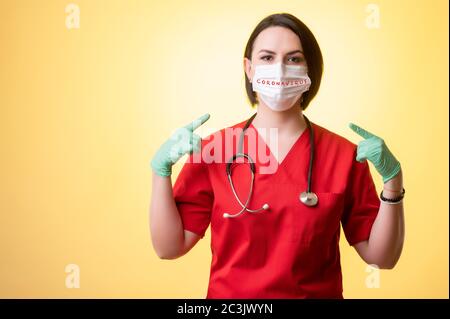 Portrait of beautiful woman doctor with stethoscope wearing red scrubs, wears a protective mask, with her fingers pointed posing on a yellow isolated Stock Photo