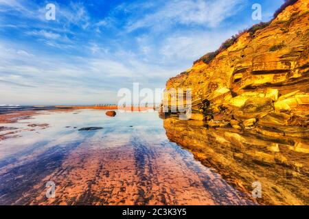 Sandstone rocks of Narrabeen headland over seabed of Narrabeen beach at low tide under warm sunrise sun. Stock Photo