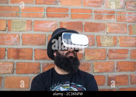 Bearded man wearing VR glasses, standing by a brick wall Stock Photo