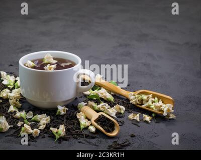 Black tea  in a white cup on a textural background, with dry tea sprinkled around and dry jasmine flowers Stock Photo