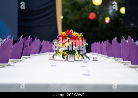 red - yellow - purple - white flower bouquet is set on the long white cover table and ready for luxury dinner in the garden field. Stock Photo
