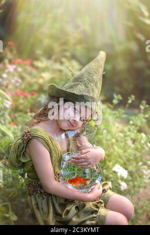 Portrait of a cute girl in a green gnome hat hugs an aquarium with a goldfish in a green garden. Stock Photo