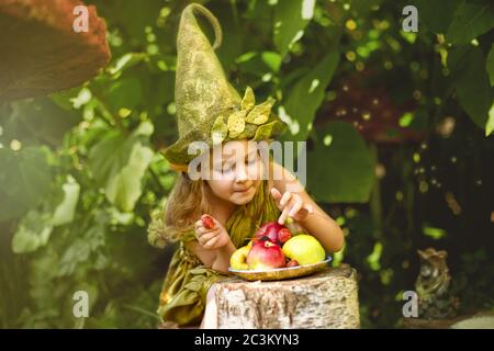 Portrait of a cute pretty girl in a gnome and costume hat in a green forest in summer. Eats berries and apples on a plate. Stock Photo