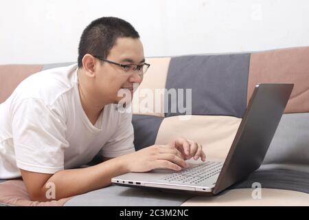 Young Asian man lying on sofa working remotely from home, Man using laptop computer having video call or buying shopping online via app while relaxing Stock Photo