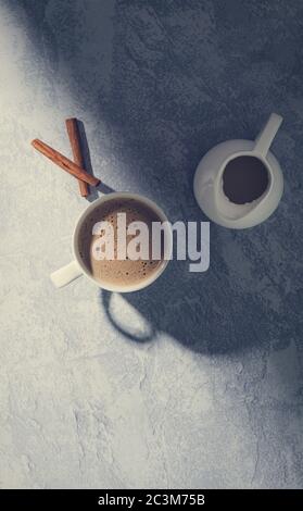 Vertical high angle shot of a cup of coffee near a white milk jug and two cinnamon near it Stock Photo