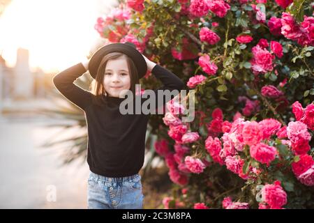 Smiling kid girl 4-5 year wearing stylish clothes posing over rose flowers at background closeup. Looking at camera. Summer season. Stock Photo