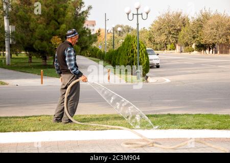Man wearing traditional uzbek hat (tubeteika) watering green lawn in Samarkand park, Uzbekistan Stock Photo