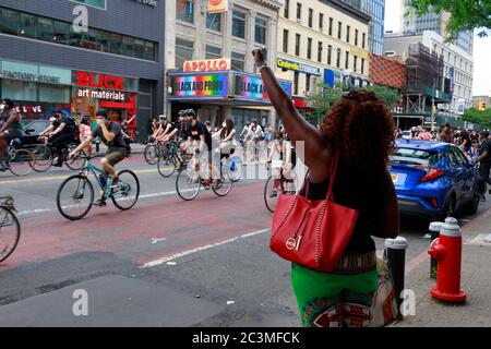 New York, NY. 20th June 2020. A woman with a raised fist stands across from the Apollo Theater with 'Black and Proud' displayed on the marquee as protesters on bicyclists ride past. The bike protest was a Black Lives Matter solidarity ride calling for justice in a recent string of American police killings: George Floyd, Breonna Taylor, and countless others. The bike ride was organized by the collective called Street Riders NYC. Several thousand people participated in the moving demonstration travelling from Times Square, Harlem, and Battery Park. June 20, 2020 Stock Photo