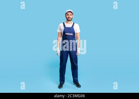 Full length handyman in overalls, cap and protective gloves standing, looking calm and positive at camera. Profession of service industry, house repai Stock Photo